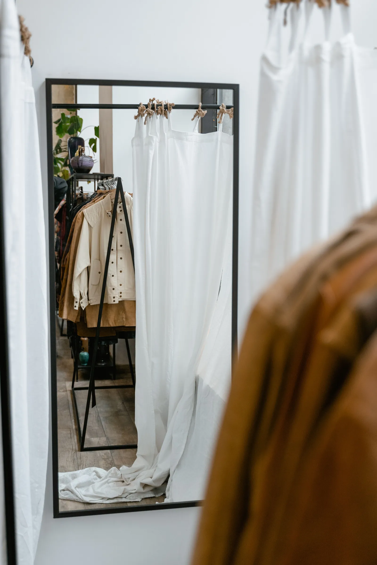 A full-length mirror in a boutique fitting area reflecting a rail of hanging garments and a white curtain, with more clothing blurred in the foreground.