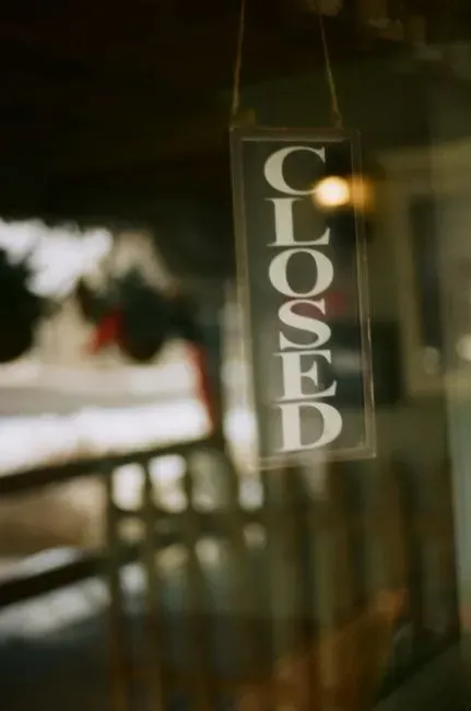 A CLOSED sign hanging in a shop window, photographed from outside through the glass so the darkened interior and a reflection of the street fall across it.