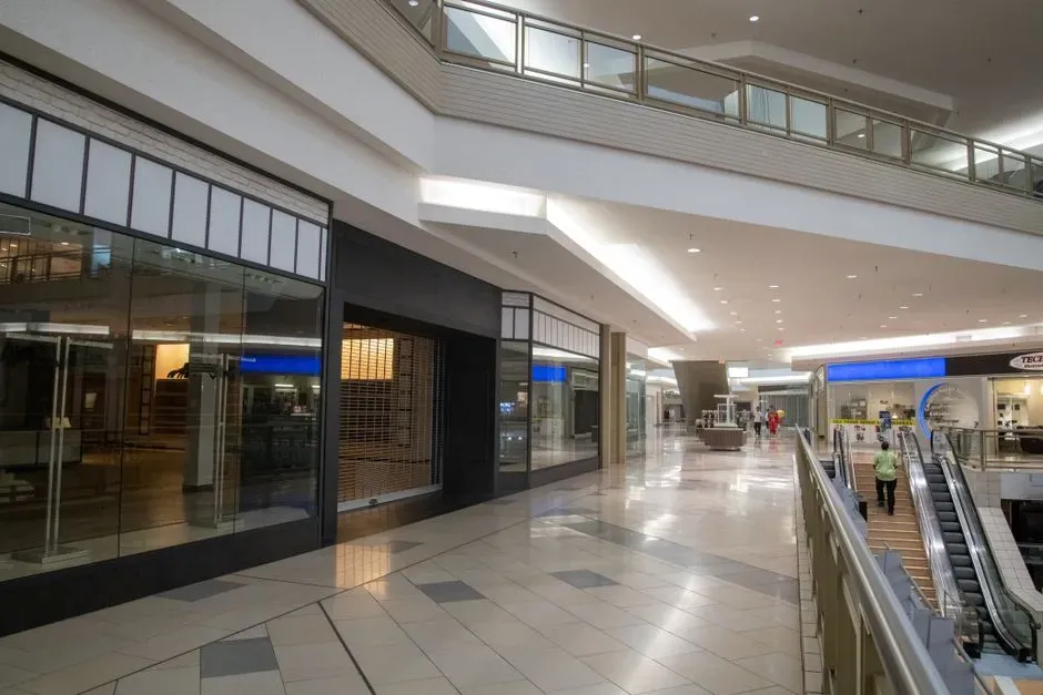 A near-empty shopping mall concourse with shuttered, unlit storefronts on both sides, an escalator on the right and only a couple of distant shoppers on the tiled floor.