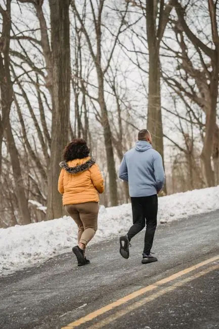 Two runners in winter jackets photographed from behind, jogging side by side along a wet road through bare woods with snow still lying on the verge.