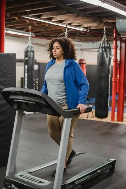 A woman in a blue jacket walking on a treadmill in a gym, red punching bags hanging on the rack behind her.