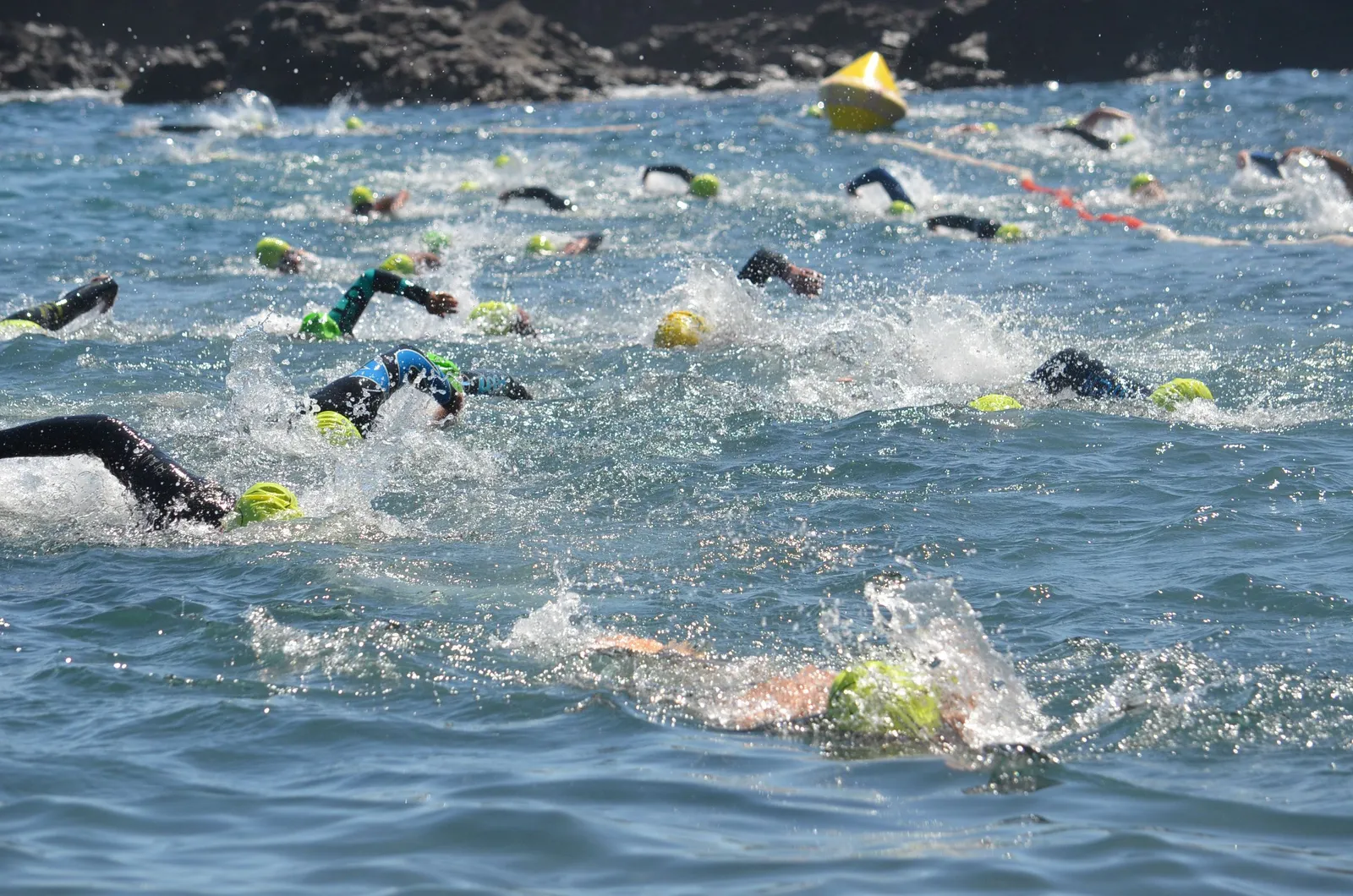 A dense pack of triathletes in wetsuits and bright swim caps churning through open water at the start of a race, a yellow marker buoy floating among them.