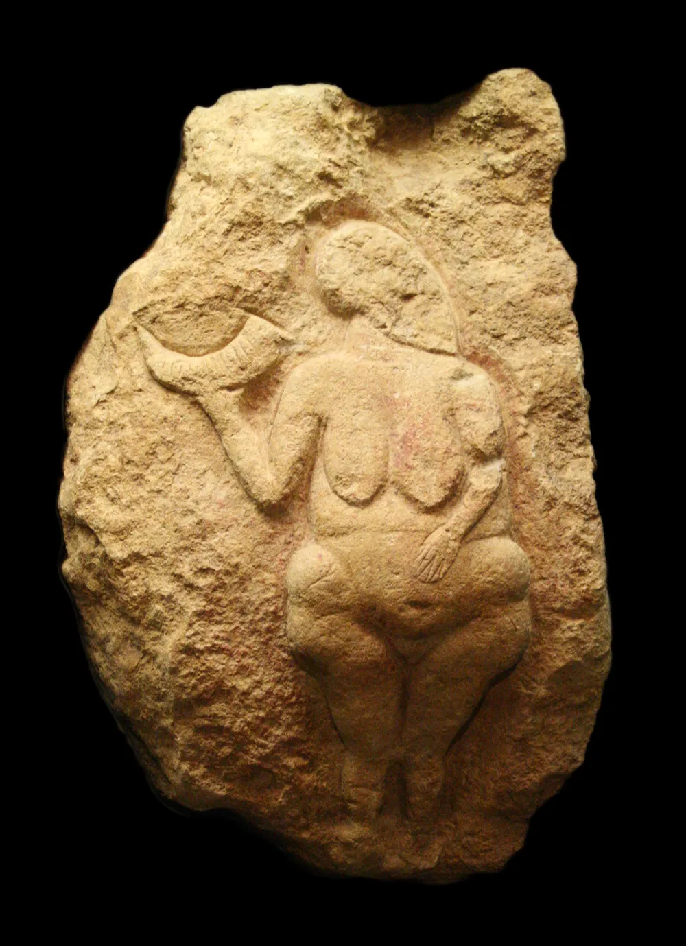 The Venus of Laussel, a limestone relief roughly 25,000 years old, carved into a block of rock: a full-bodied woman in profile-turned-frontal pose, one hand on her belly and the other raising a notched bison horn above her shoulder.