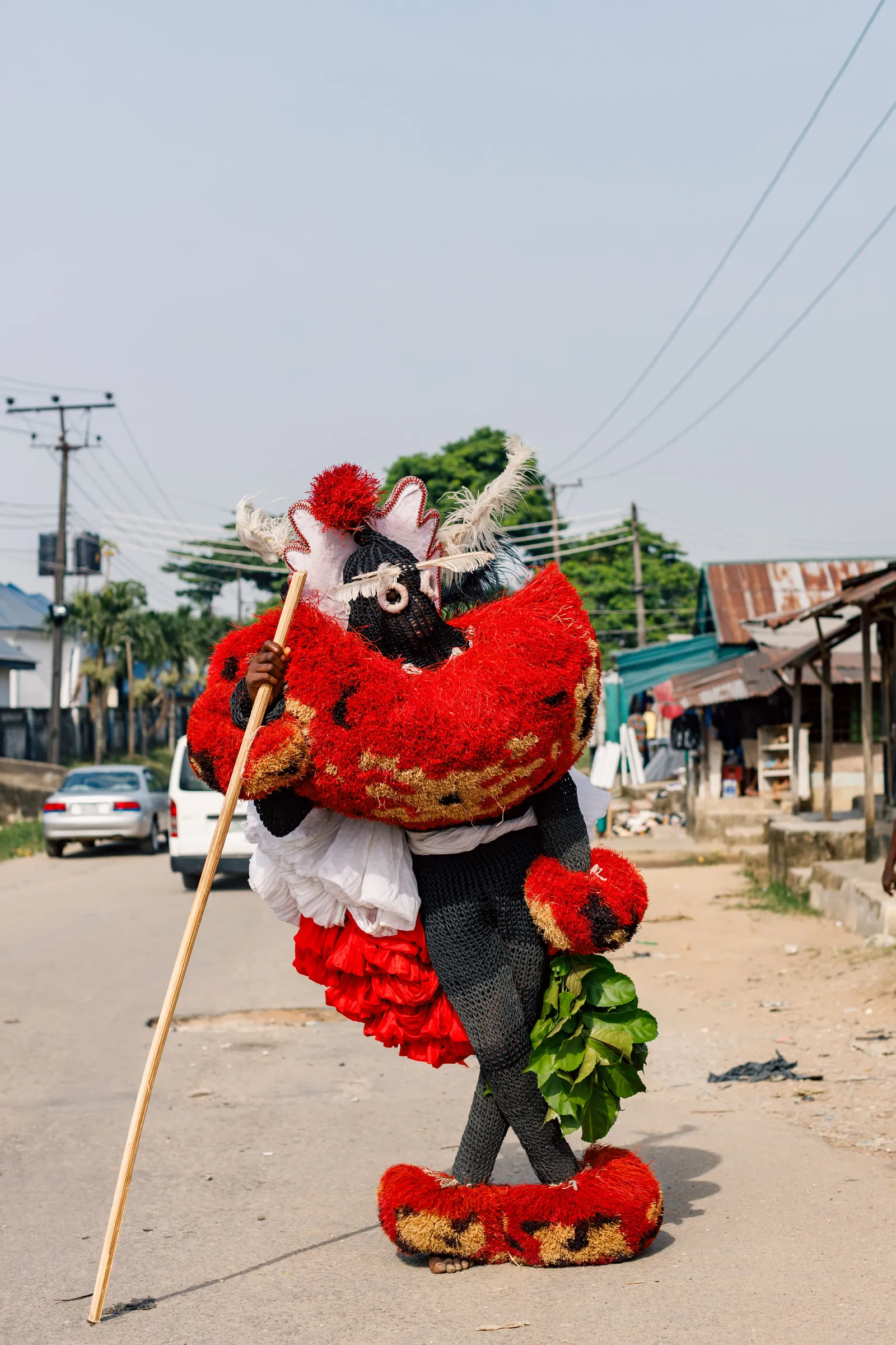 A masquerade performer in Calabar, Nigeria, walking a sandy street in a full costume of dense red and green raffia with a carved headpiece and matching anklets, carrying a long staff. The costume covers the wearer entirely.