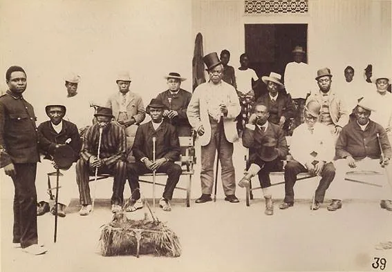An early photograph of Obong Orok Edem-Odo seated with the chiefs of Old Calabar, around twenty men arranged in rows outside a building, most in suits and hats, several holding carved staffs, with ceremonial fans and a bundle of regalia set on the ground in front of them.