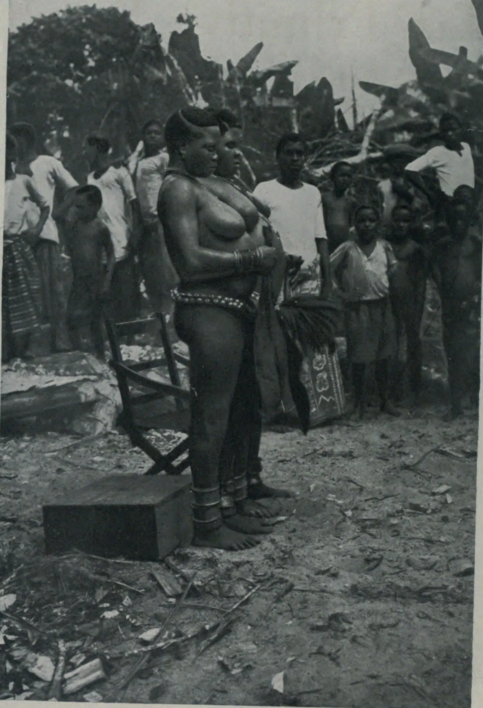 A 1915 photograph captioned Fatting-House Women. A full-figured young woman stands in profile in an open clearing, wearing strings of beads at her waist, heavy brass bracelets and thick brass anklets, her hair dressed in a high crested style. Behind her a crowd of townspeople — men, women and children — stands watching.