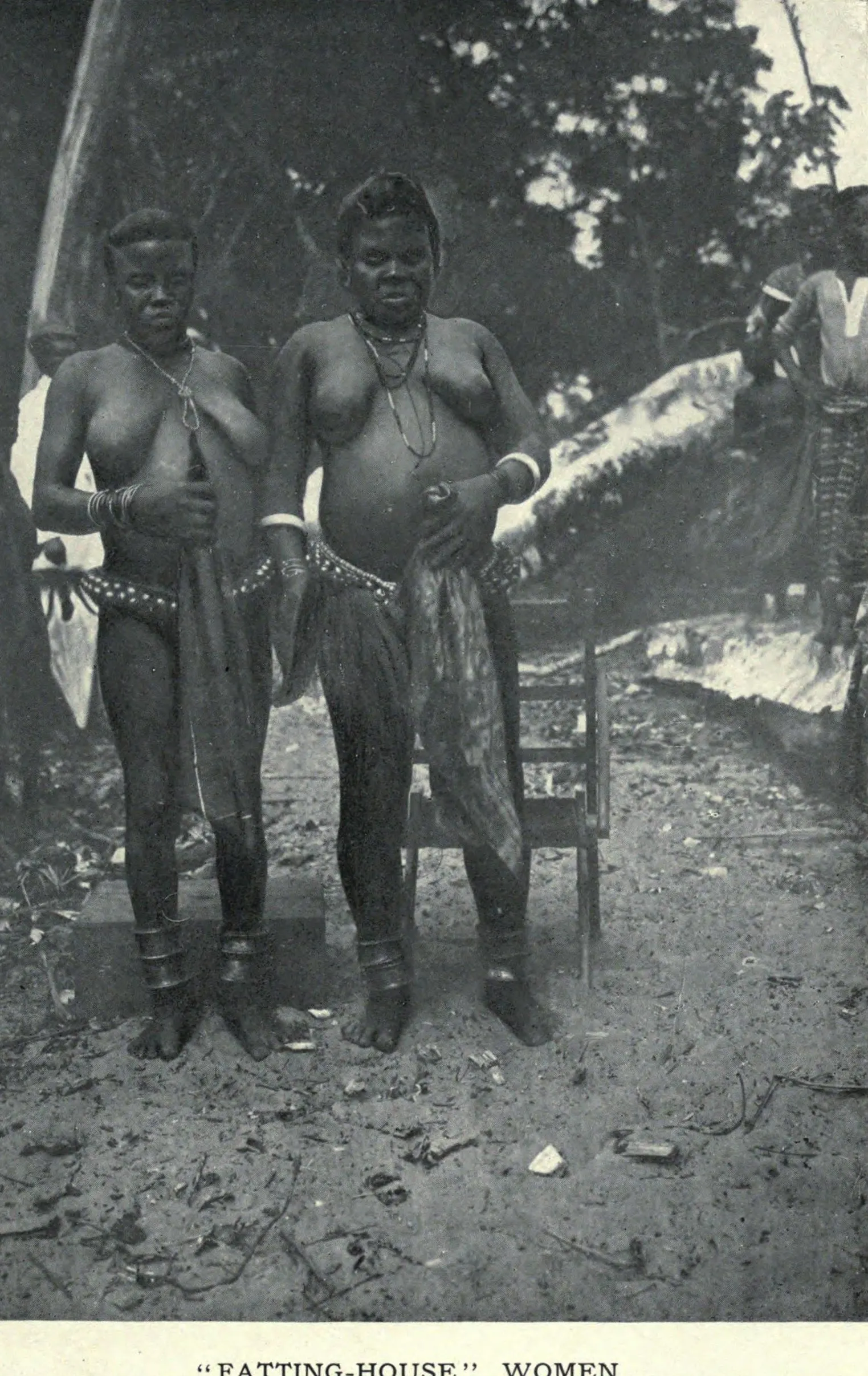 A 1915 photograph captioned Fatting-House Women. Two women stand side by side in a clearing, both wearing beaded waist strings, brass bracelets and heavy brass anklets, each holding a length of patterned cloth. A felled palm and another figure are visible behind them.