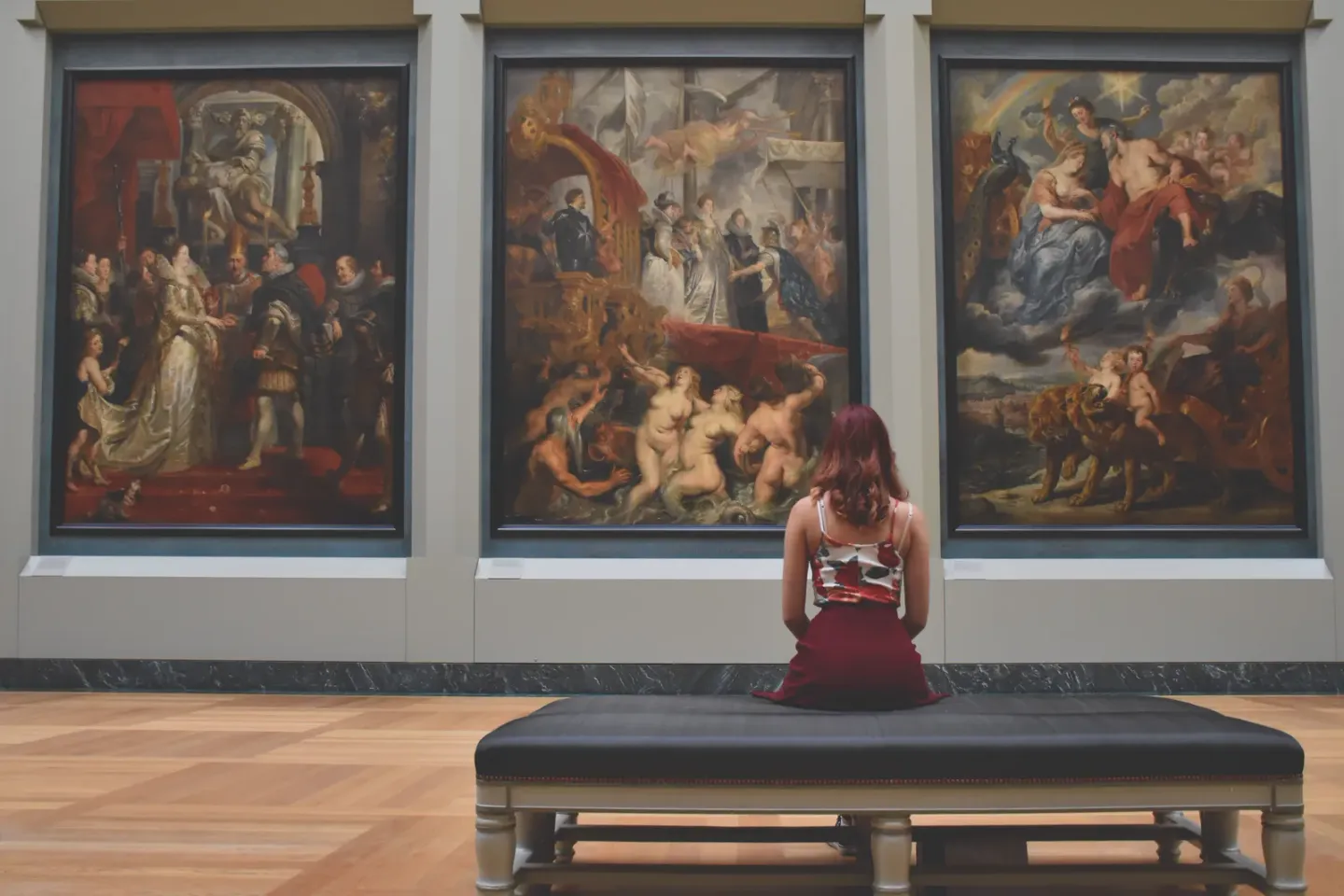 A visitor sitting alone on a bench in a museum gallery, viewed from behind, facing three large gilt-framed old master paintings hung on a pale wall.