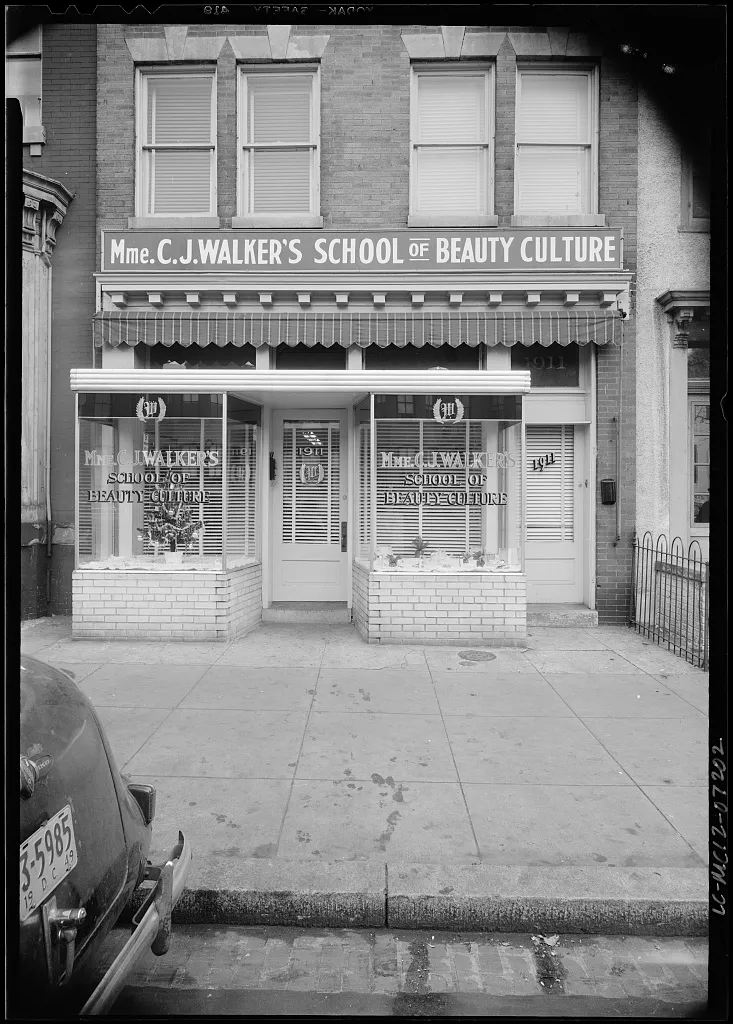 Storefront of Madam C. J. Walker’s School of Beauty Culture photographed in 1949: a painted sign reading MME. C. J. WALKER’S SCHOOL OF BEAUTY CULTURE runs above a shopfront with a striped awning and lettered display windows, a period car parked at the kerb.