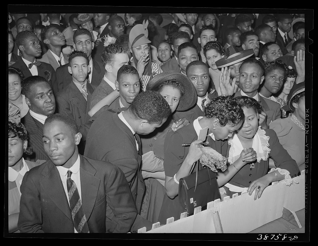 A dense crowd of Black men and women in suits, hats and evening dress pressed together at a ballroom railing in 1941 - some watching the floor, some talking, one woman leaning in to speak to another over the noise.