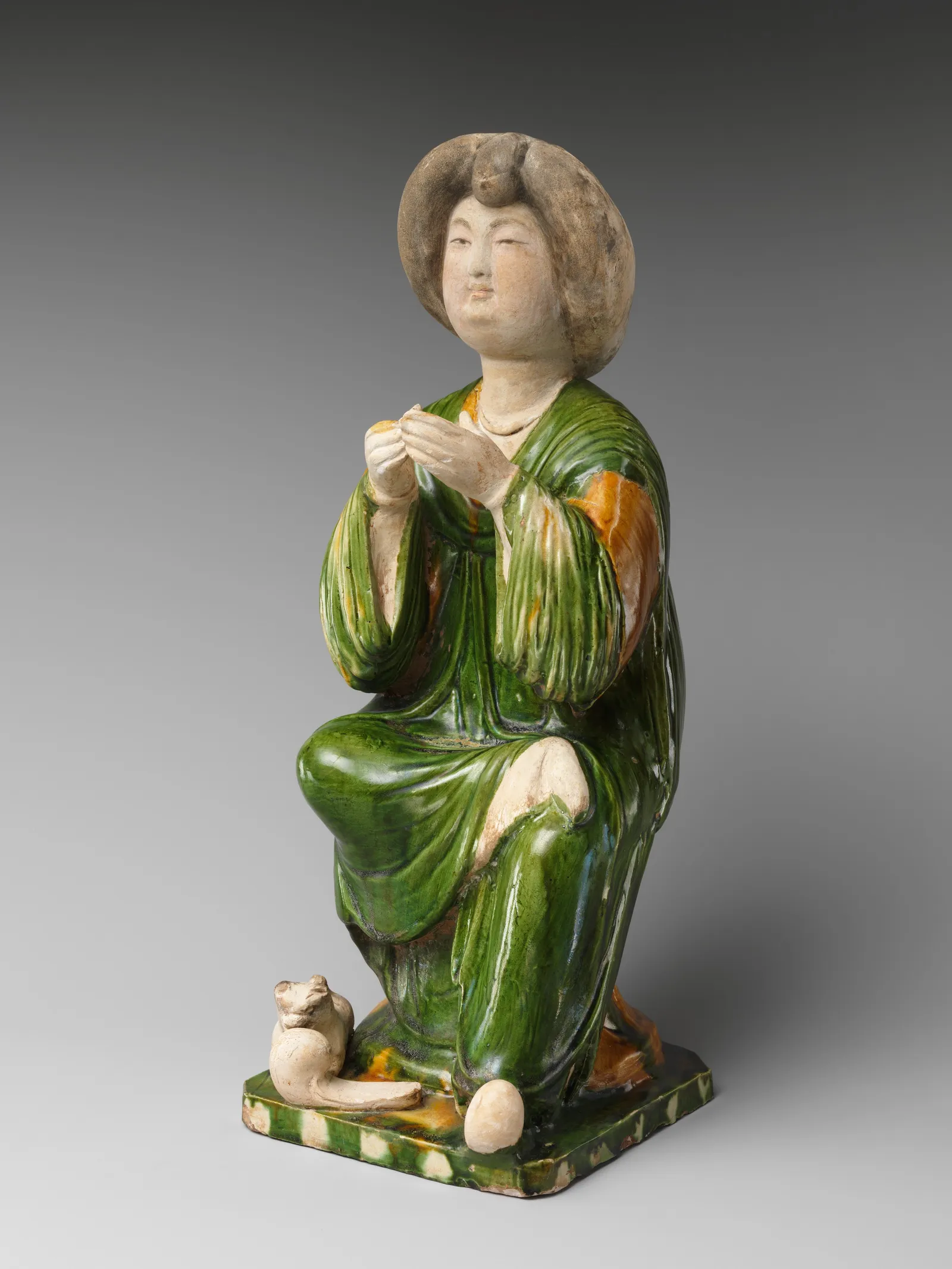 An eighth-century Chinese tomb figurine of a seated court lady in earthenware with a green, amber and cream sancai glaze. She sits with her hands raised together at her chest, her face round and full and her body broad beneath heavy draped robes — the build Tang sculptors treated as the court ideal.