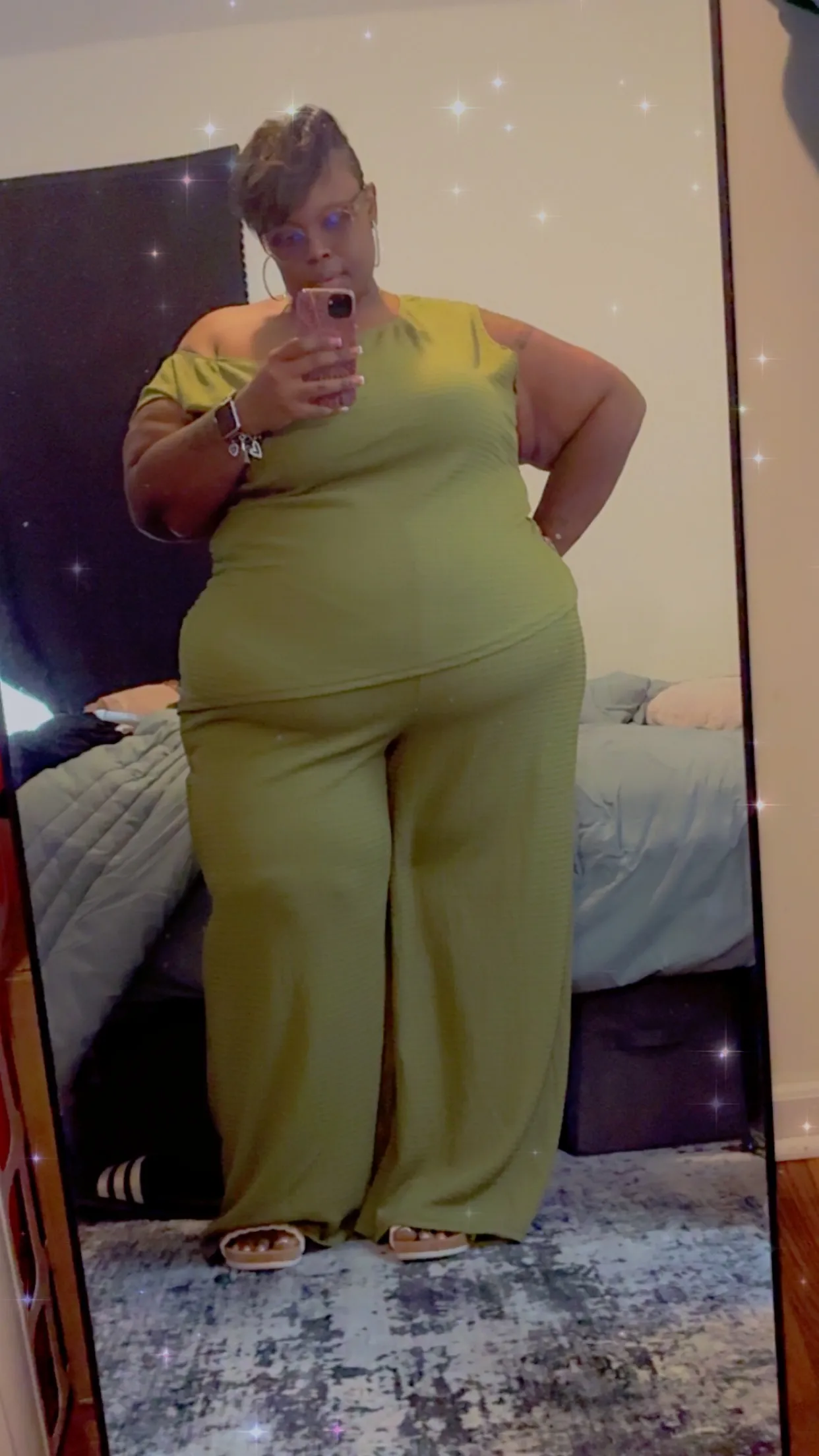 Yvonne taking a mirror selfie in an olive-green off-shoulder jumpsuit in her bedroom.