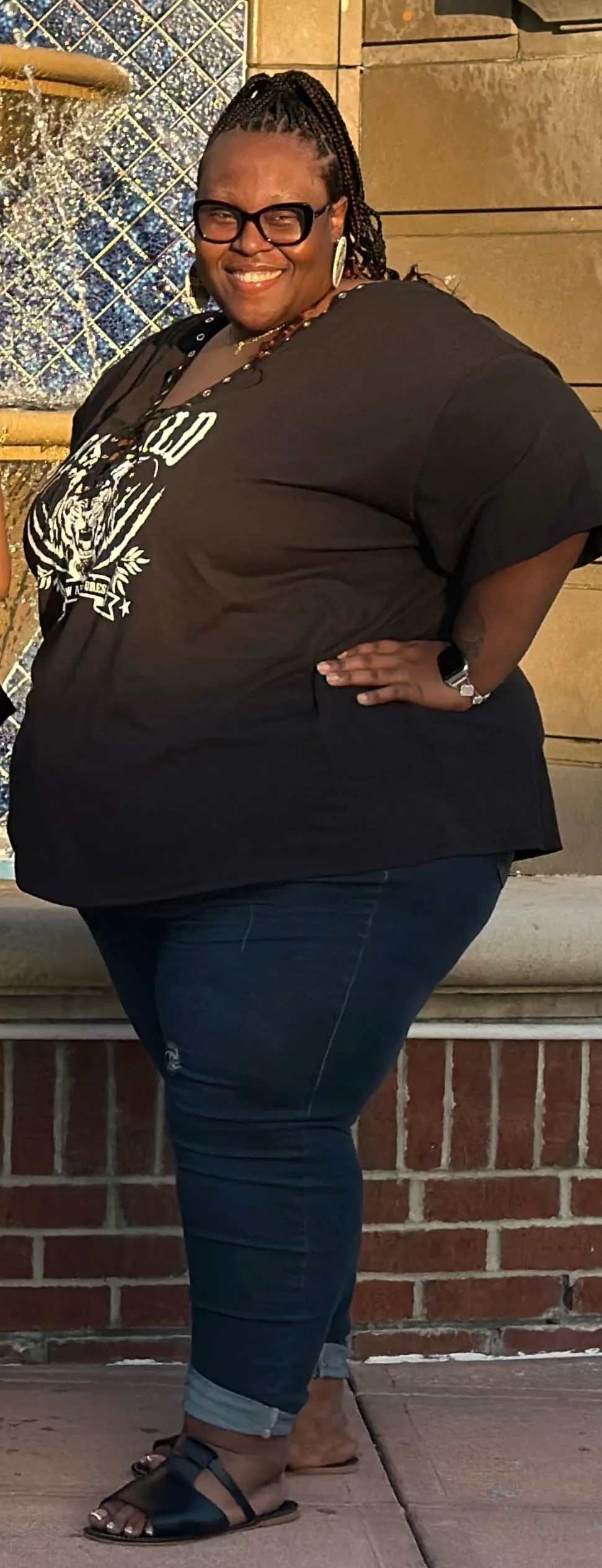 Yvonne smiling outdoors by a fountain, wearing a dark brown t-shirt with a white tiger graphic and jeans.