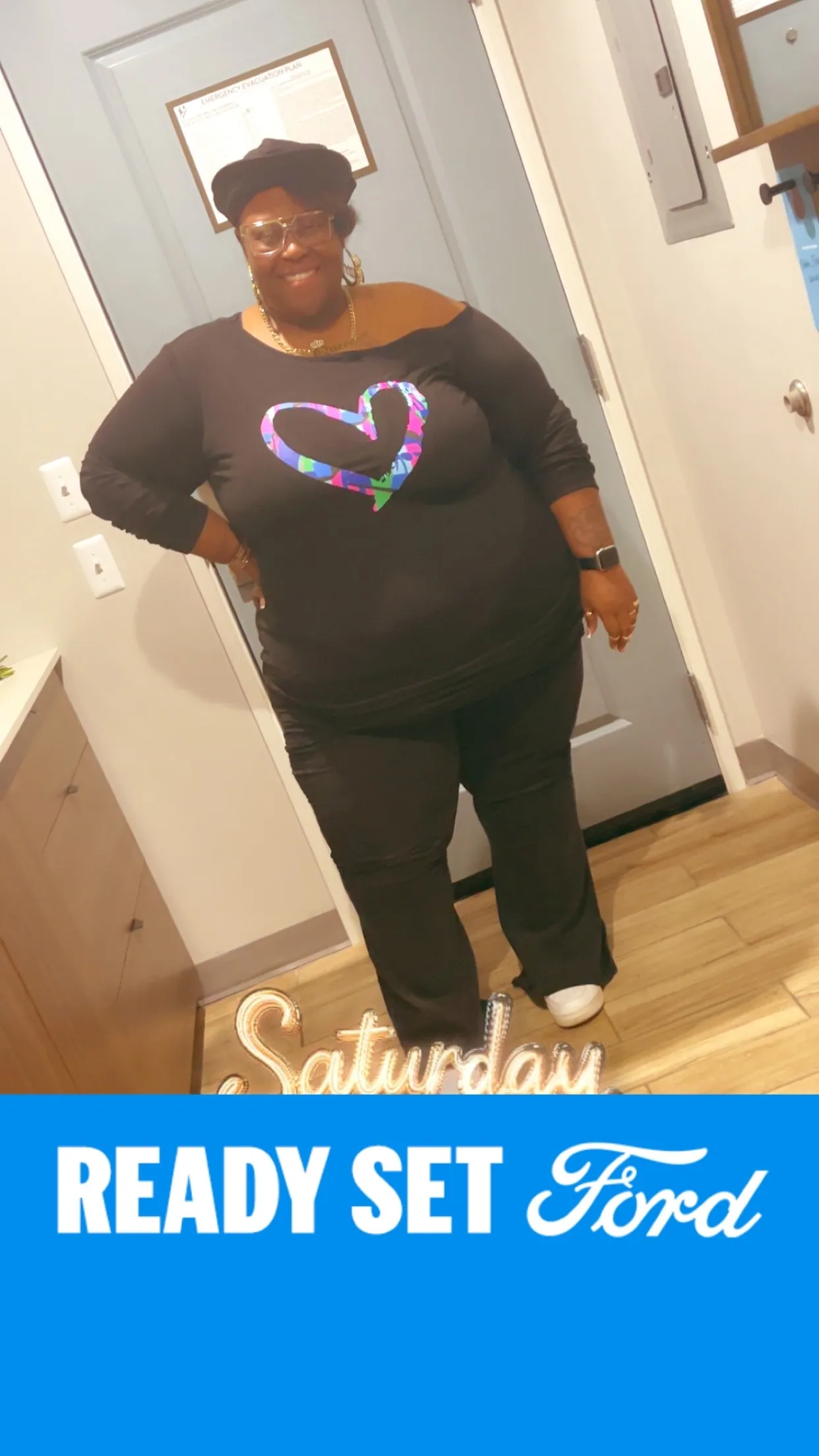 Yvonne smiling in a black top with a colorful heart graphic and a black hat, indoors.