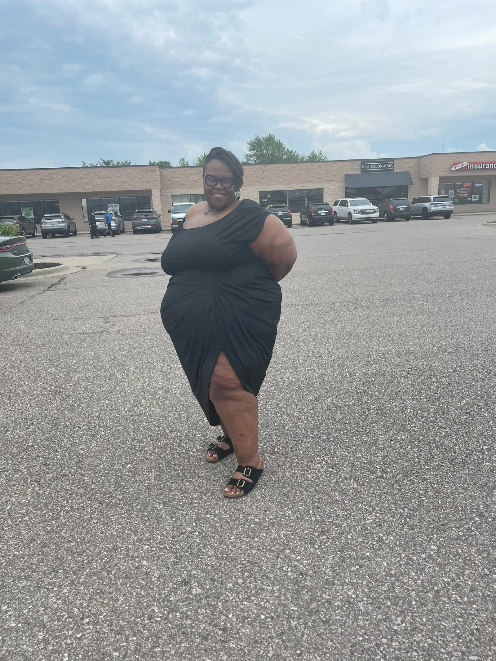 Yvonne posing in a black one-shoulder draped dress in an outdoor parking lot.